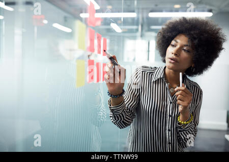 donna d'affari che guarda le note adesive sulla parete in vetro in ufficio Foto Stock