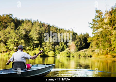 Vista posteriore dell'uomo la pesca mentre sedendo nel canotto sul lago contro alberi e cielo Foto Stock