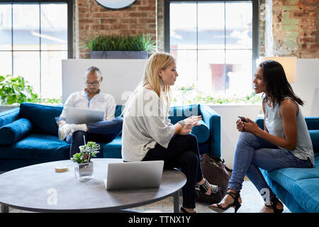 donne d'affari che discutono e colleghi maschi che lavorano su un computer portatile mentre sono seduti sul divano in ufficio Foto Stock