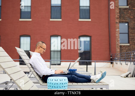 Vista laterale dell'uomo d'affari con computer portatile mentre è seduto sulla sedia a sdraio sulla terrazza edificio Foto Stock