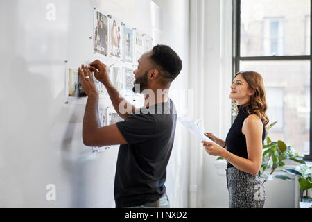uomo d'affari che attacca le stampe fotografiche sulla bacheca mentre sorride una collega donna che gli sta accanto in ufficio Foto Stock