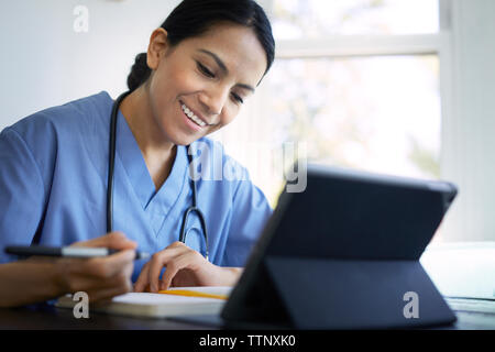 Donna sorridente medico iscritto sul diario mentre si lavora alla scrivania in ospedale Foto Stock