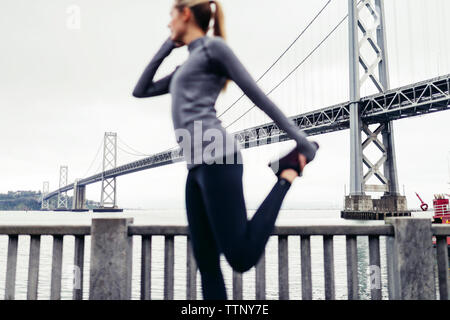 Vista laterale di atleta femminile allungamento della gamba sul sentiero con Oakland Bay Bridge in background Foto Stock