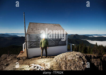 Uomo che indossa testa lampada mentre in piedi contro la cabina in legno sulla montagna al crepuscolo Foto Stock