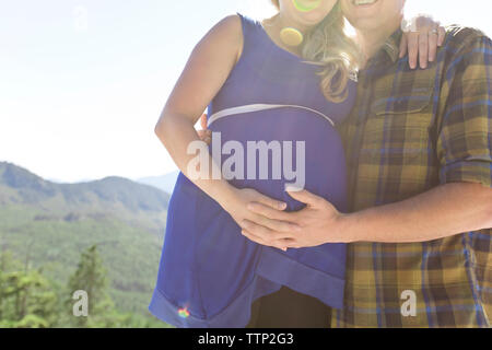 Toccare uomo donna incinta lo stomaco mentre in piedi contro montagne durante la giornata di sole Foto Stock