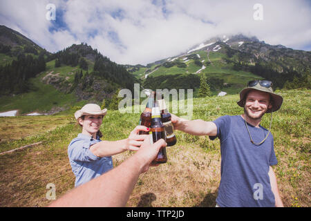 Happy amici tostare le bottiglie di birra sul campo contro le montagne e il cielo nuvoloso Foto Stock