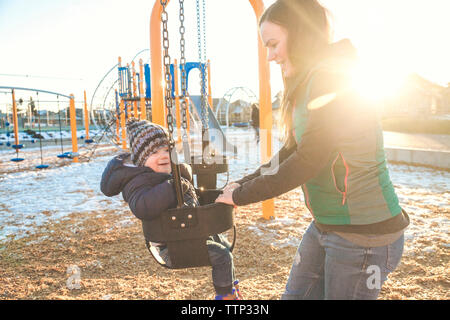 Madre guardando il ragazzo giocando su swing durante il periodo invernale Foto Stock