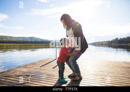 Vista laterale del figlio amorevole baciare sulla gravidanza della madre ventre sopra il lago contro sky durante la giornata di sole Foto Stock