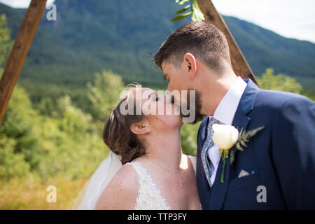 Sposa giovane kissing mentre in piedi contro la montagna nella cerimonia di nozze Foto Stock