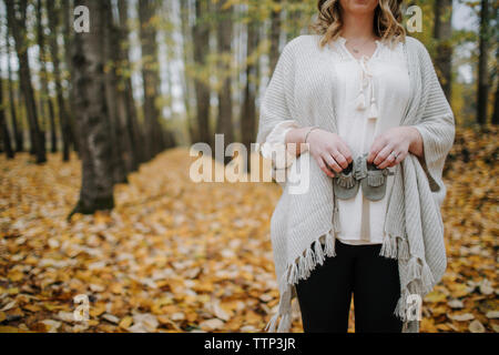 Sezione mediana della donna incinta holding baby babbucce permanente, mentre nella foresta durante l'autunno Foto Stock