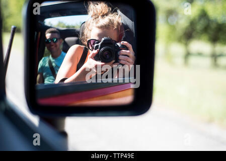 Donna Fotografa riflettendo sul lato specchietto retrovisore mentre si viaggia con un amico in fuoristrada Foto Stock