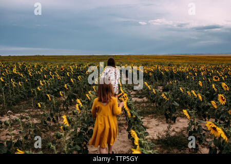 Vista posteriore di suore a piedi nella fattoria di girasole contro il cielo nuvoloso Foto Stock