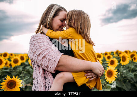 Madre abbracciando la figlia mentre in piedi nella fattoria di girasole contro sky Foto Stock