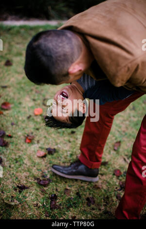 Elevato angolo di visione del padre giocoso che porta felice figlio mentre si sta in piedi sul campo erboso in Balboa Park Foto Stock