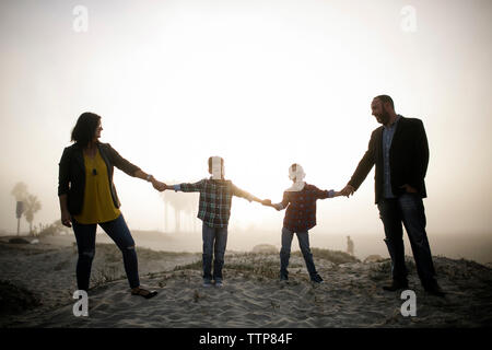 Felice famiglia silhouette tenendo le mani mentre si sta in piedi in spiaggia contro il cielo durante la nebbia meteo Foto Stock