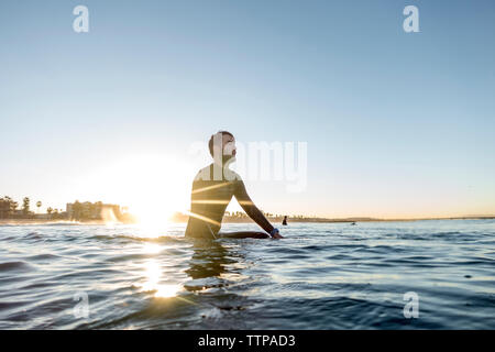 Riflessivo surfer seduto sulla tavola da surf in mare contro il cielo chiaro durante il tramonto Foto Stock
