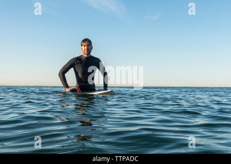 Surfista maschile seduto sulla tavola da surf in mare contro il cielo chiaro Foto Stock