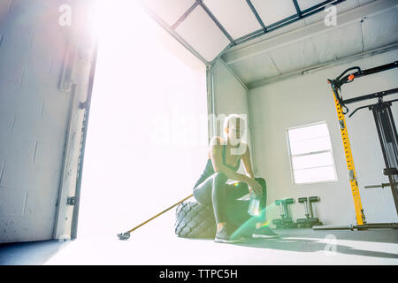 Stanco donna seduta sul pneumatico mentre il contenimento dell'acqua in bottiglia luminosa palestra Foto Stock