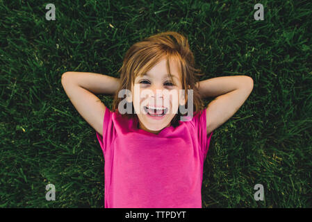 Ritratto di overhead di allegro ragazza con le mani dietro la testa mentre giaceva sul campo erboso Foto Stock