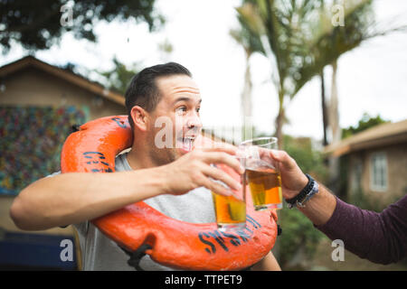 Immagine ritagliata della mano la birra di tostatura di vetro con felice amico mentre in piedi in cortile Foto Stock