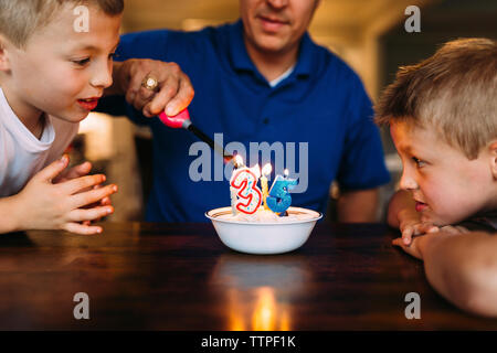 Curioso fratelli guardando padre illuminazione candele di compleanno con leggera sul tavolo a casa Foto Stock