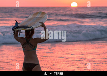 Vista posteriore della donna che la porta con la tavola da surf in piedi contro il mare Foto Stock