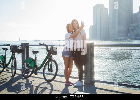 Coppia felice tenendo selfie attraverso il telefono cellulare mentre in piedi in bici durante la giornata di sole Foto Stock