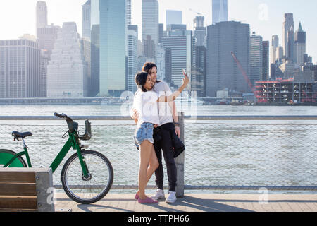 Giovane tenendo selfie sul telefono cellulare mentre in piedi da biciclette contro il paesaggio urbano Foto Stock