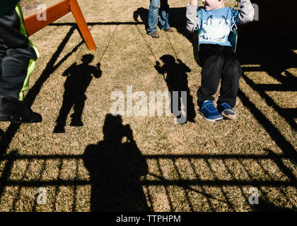 Ombra di figli oscillazioni durante il padre permanente al parco giochi Foto Stock