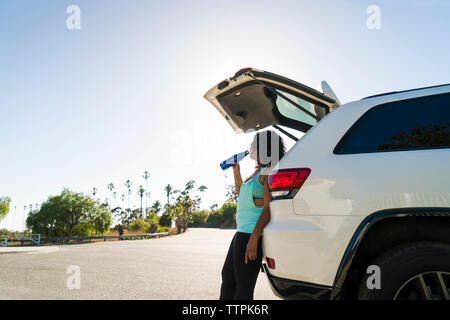 Vista laterale della donna acqua potabile mentre appoggiata contro l'auto Foto Stock