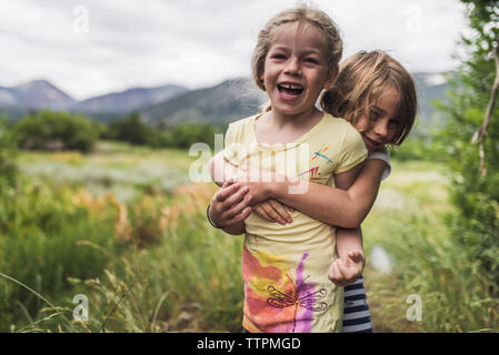 Ritratto di felice carino sorelle abbracciando mentre si sta in piedi sul paesaggio contro il cielo nuvoloso in foresta Foto Stock