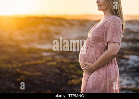 Sezione mediana della donna incinta con le mani sul ventre in piedi in spiaggia contro il cielo chiaro durante il tramonto Foto Stock