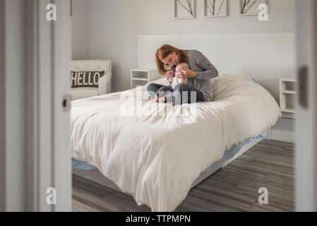 Felice madre giocando con il simpatico figlio seduti sul letto contro la parete a casa visto attraverso la porta Foto Stock