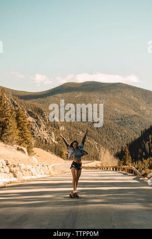 Lunghezza completa di spensierata giovane donna a braccia alzate con lo skateboard su strada contro le montagne Foto Stock