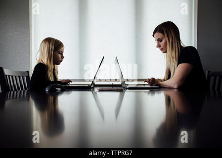 Vista laterale della madre e figlia utilizzando computer portatili sul tavolo mentre è seduto a casa Foto Stock
