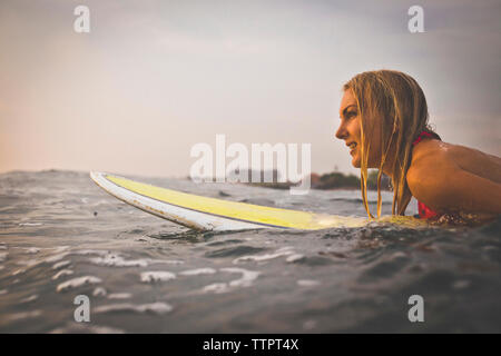 Donna che guarda lontano mentre giaceva sulla tavola da surf in mare contro sky Foto Stock