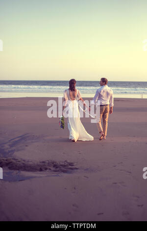 Vista posteriore della sposa giovane tenendo le mani mentre passeggiate in spiaggia contro il cielo chiaro durante il tramonto Foto Stock