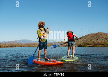 Lunghezza completa paddleboarding amici sul lago contro il cielo blu chiaro Foto Stock