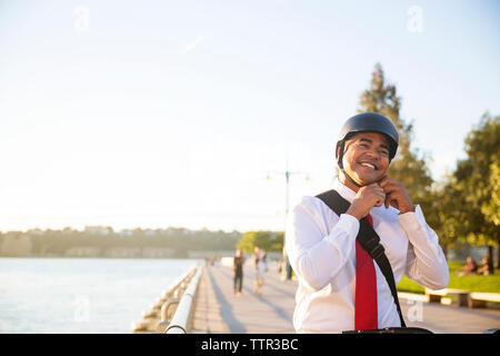 Happy businessman indossando il casco in bicicletta mentre in piedi contro il cielo Foto Stock