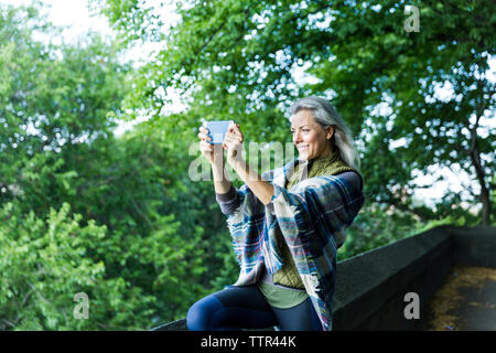 Donna fotografando attraverso smart phone mentre si sta in piedi sul sentiero contro i rami Foto Stock