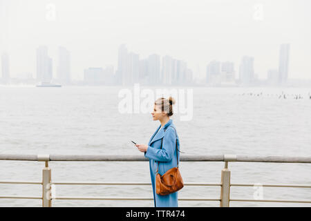 Vista laterale della giovane donna con portamonete indossa trench coat tenendo premuto smart phone dalla ringhiera e fiume in città Foto Stock