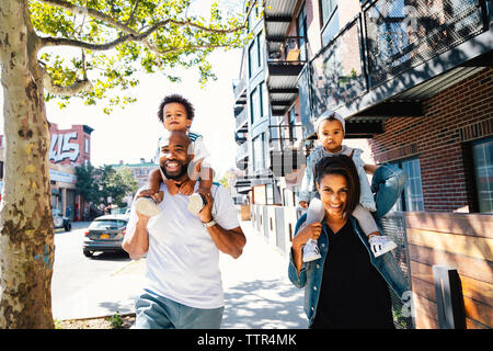 Ritratto di genitori felici il trasporto di bambini su spalle mentre camminando sul marciapiede in città Foto Stock