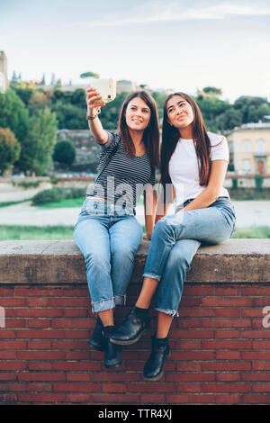 Lunghezza completa di felice amici di sesso femminile prendendo selfie con fotocamera istantanea mentre è seduto sulla parete di ritegno in posizione di parcheggio Foto Stock