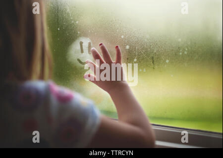 Immagine ritagliata della ragazza rendendo antropomorfi Smiley face sulla finestra bagnata a casa durante la stagione delle piogge Foto Stock