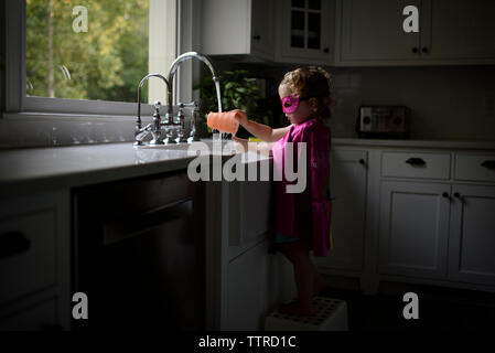 Vista laterale della ragazza di indossare il costume del supereroe di lavarsi le mani nel lavello da cucina mentre in piedi a casa Foto Stock