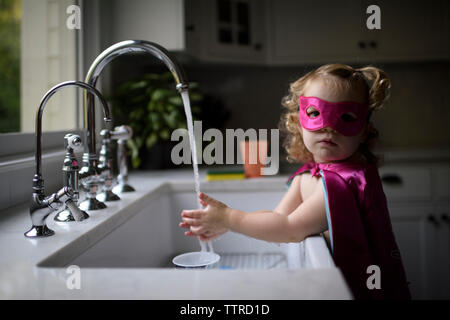 Ritratto di una ragazza indossando il costume del supereroe di lavarsi le mani nel lavello da cucina mentre in piedi a casa Foto Stock