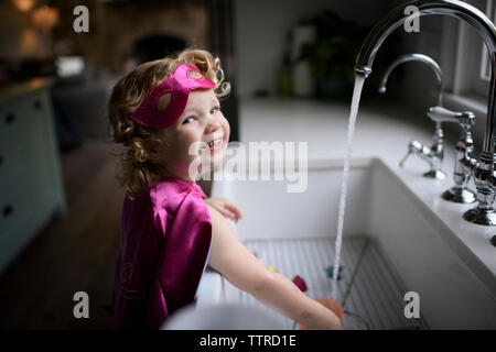 Ritratto di ragazza sorridente indossando il costume del supereroe di lavarsi le mani nel lavello da cucina mentre in piedi a casa Foto Stock