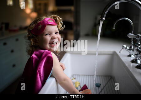 Ritratto di ragazza sorridente indossando il costume del supereroe di lavarsi le mani nel lavello da cucina a casa Foto Stock