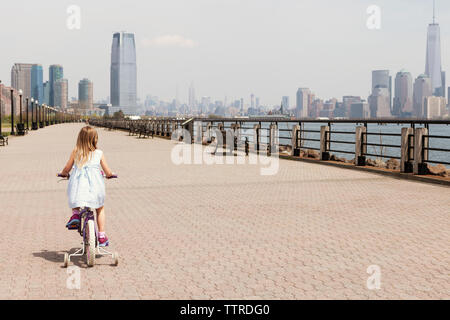 Vista posteriore di una ragazza in bicicletta sul lungomare con vista sullo skyline in background Foto Stock