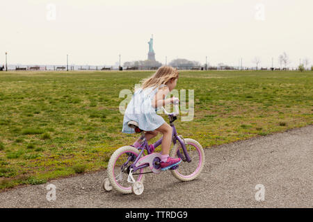 Vista laterale della ragazza Bicicletta Equitazione sulla strada dal campo con la Statua della Libertà in background Foto Stock